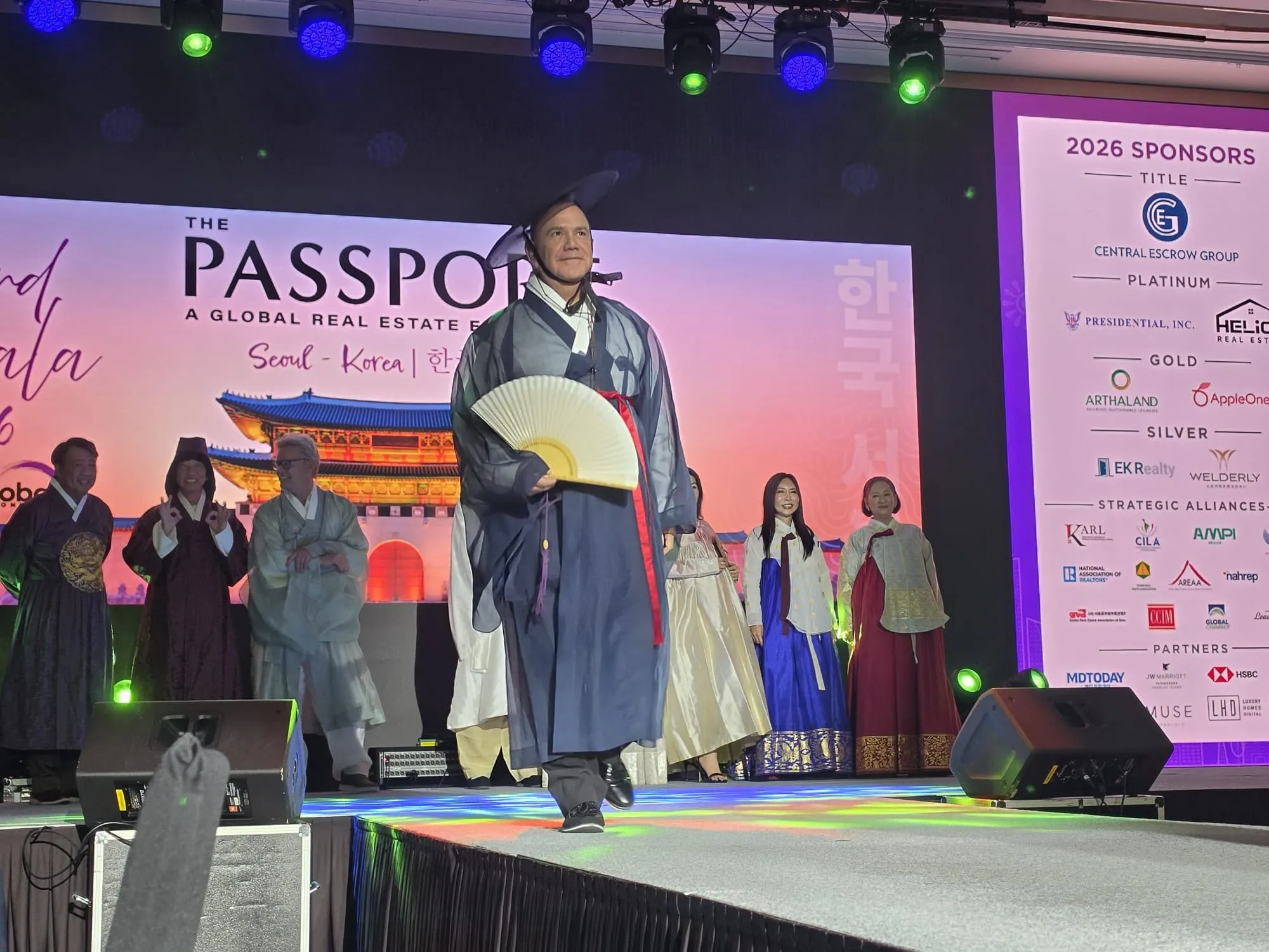 Kiko walking across the stage at The Passport gala in Seoul, in a blue traditional Korean robe and hat, holding a fan